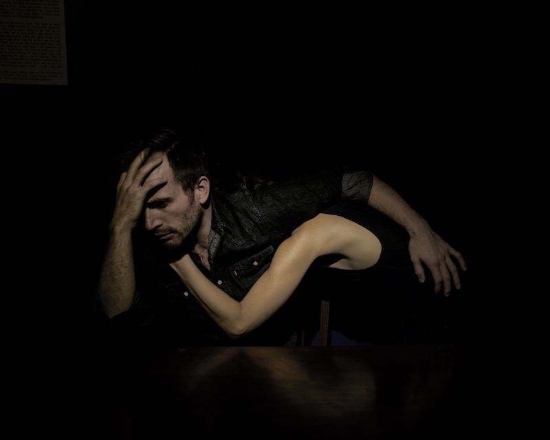 A man in denim shirt holds his head as if he's in distress. A woman embraces him. Her face hidden from the camera. All we see is her bare arm wrapped around his chest.